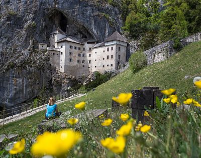 Predjamski grad, pred njim travnik s cvetičimi zlaticami, ženska v modri majici sedi na klopci, gleda grad