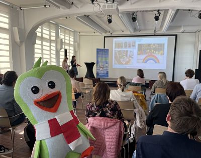 A plush Green Penguin mascot is placed in the centre, while participants watch the presentation on a screen in the background.