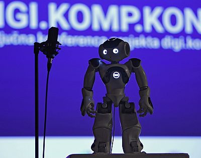 A robot stands on a stage with a microphone next to it. Behind the robot is a large screen with a presentation.