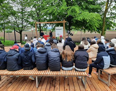 dijaki in drugi udeleženci sedijo na klopeh na zelenici in poslušajo govor dveh dijakov