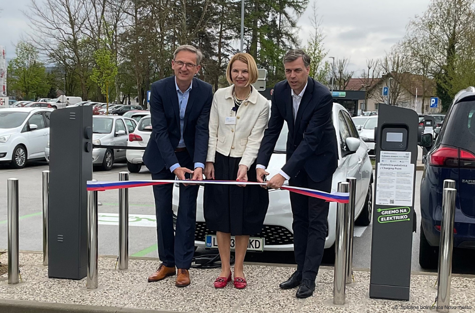 A man, a woman and another man stand in a line in the parking holding a ribbon in the colours of the Slovenian flag, which is stretched between two charging stations for electric vehicles, to be cut.