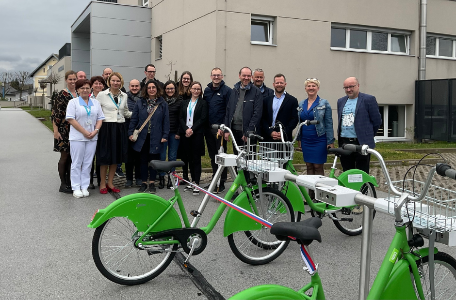 A group of people stands in front of the building and behind the bike shed, with a ribbon in the colours of the Slovenian flag stretched between two bicycles, ready for the opening ceremony.