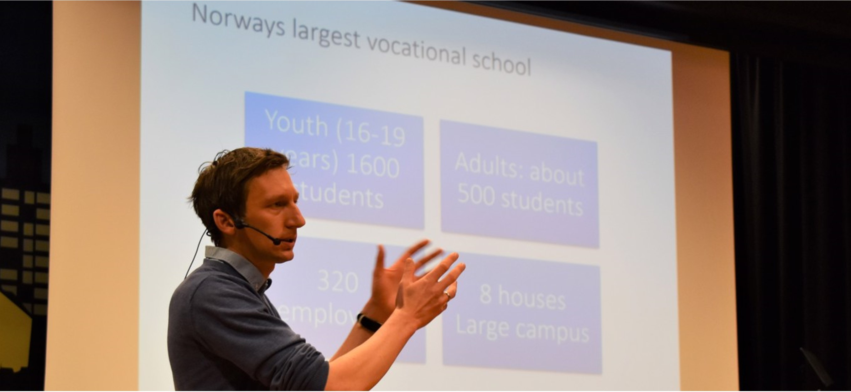 A man lectures with a microphone on his head and his arms outstretched in front of a screen on which a presentation is being shown. 