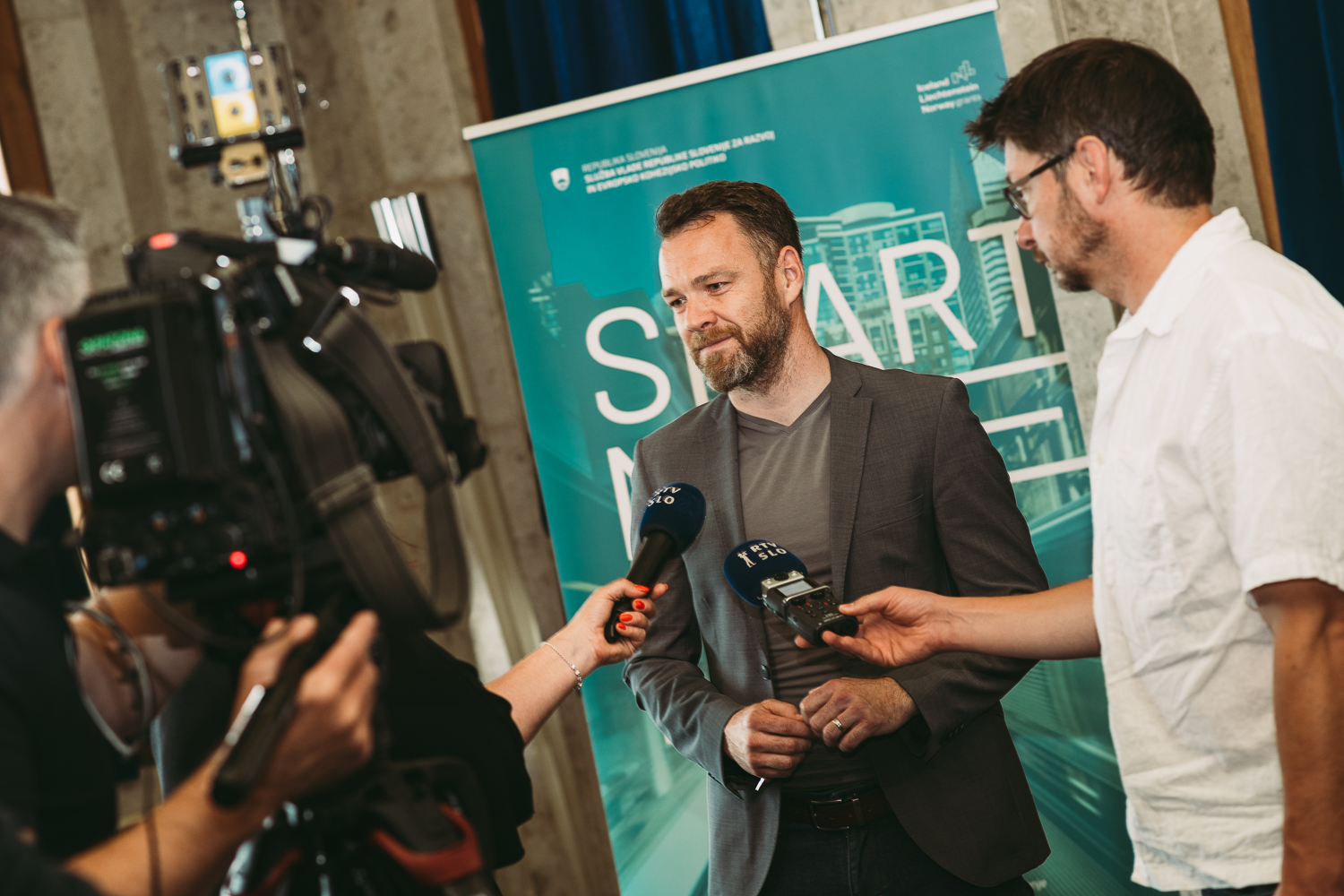 Two journalists, holding microphones in their hands, are interviewing a man in front of a promotional poster, while a cameraman is recording with a camera.