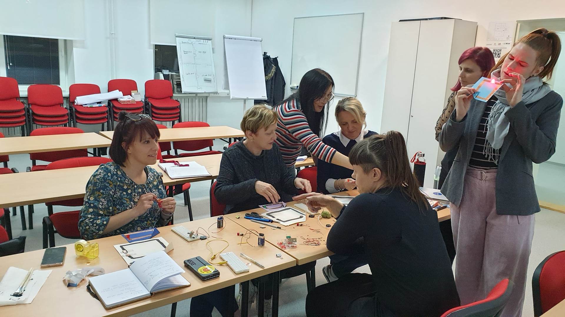 A group of women around a table, on the table are cables, chips, a notebook, tape.