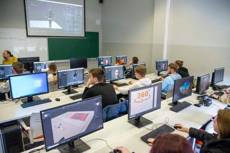 Students at their desks, each with a screen on their desk, look at a screen projecting the workings of a computer programme.