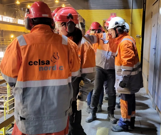 A group of people wearing protective clothing, goggles, headphones and helmets look down on the production hall from a balcony. 