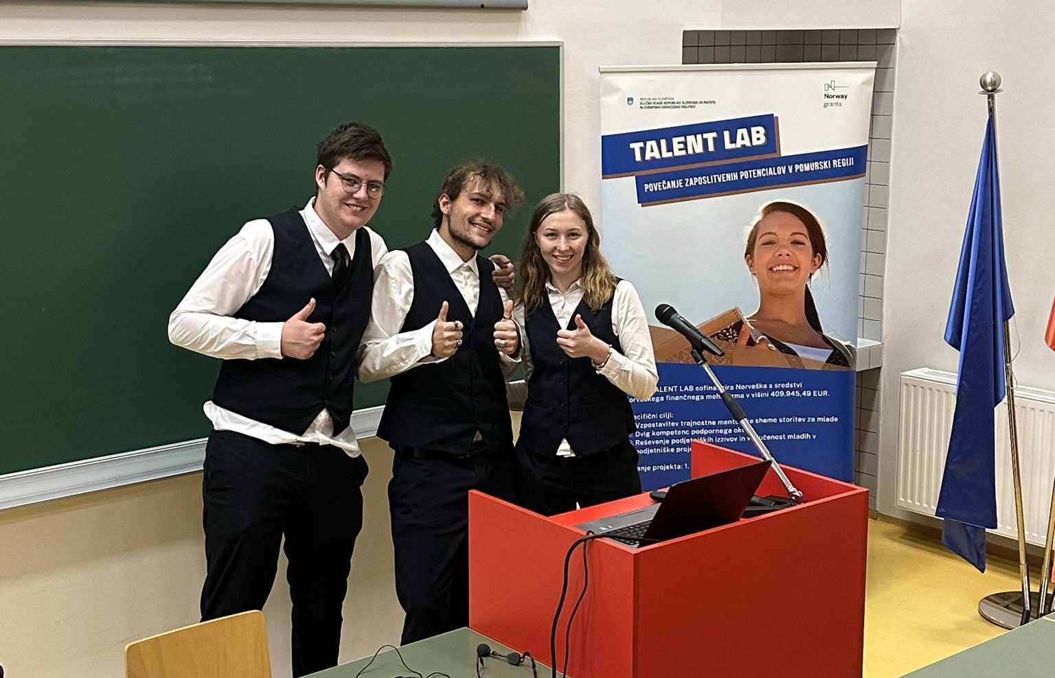 Trije dijaki za govorniškim odrom se smehljajo in kažejo stegnjen palec, zadaj je telena tabla in reklamno stojalo za projekt Talent LAB