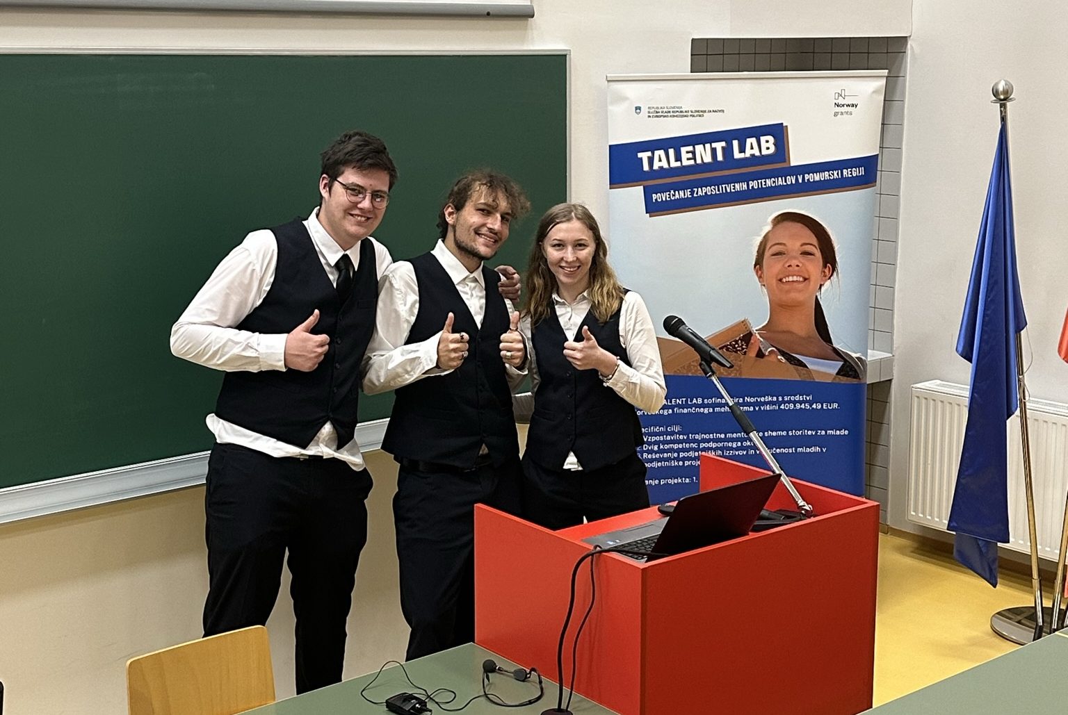 Three students at the podium smile and give a thumbs-up, behind them is a TV board and a promotional stand for the Talent LAB project.