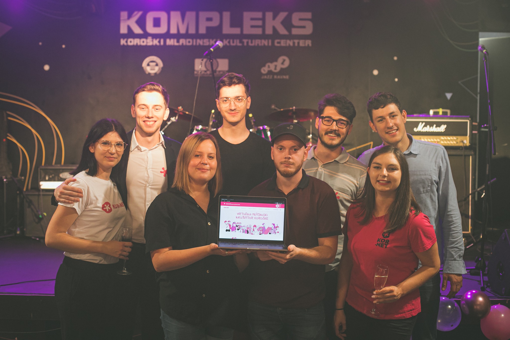 A group of people in front of a music stage, two of them holding a laptop with the Virtual Youth Incubator of Carinthia website open