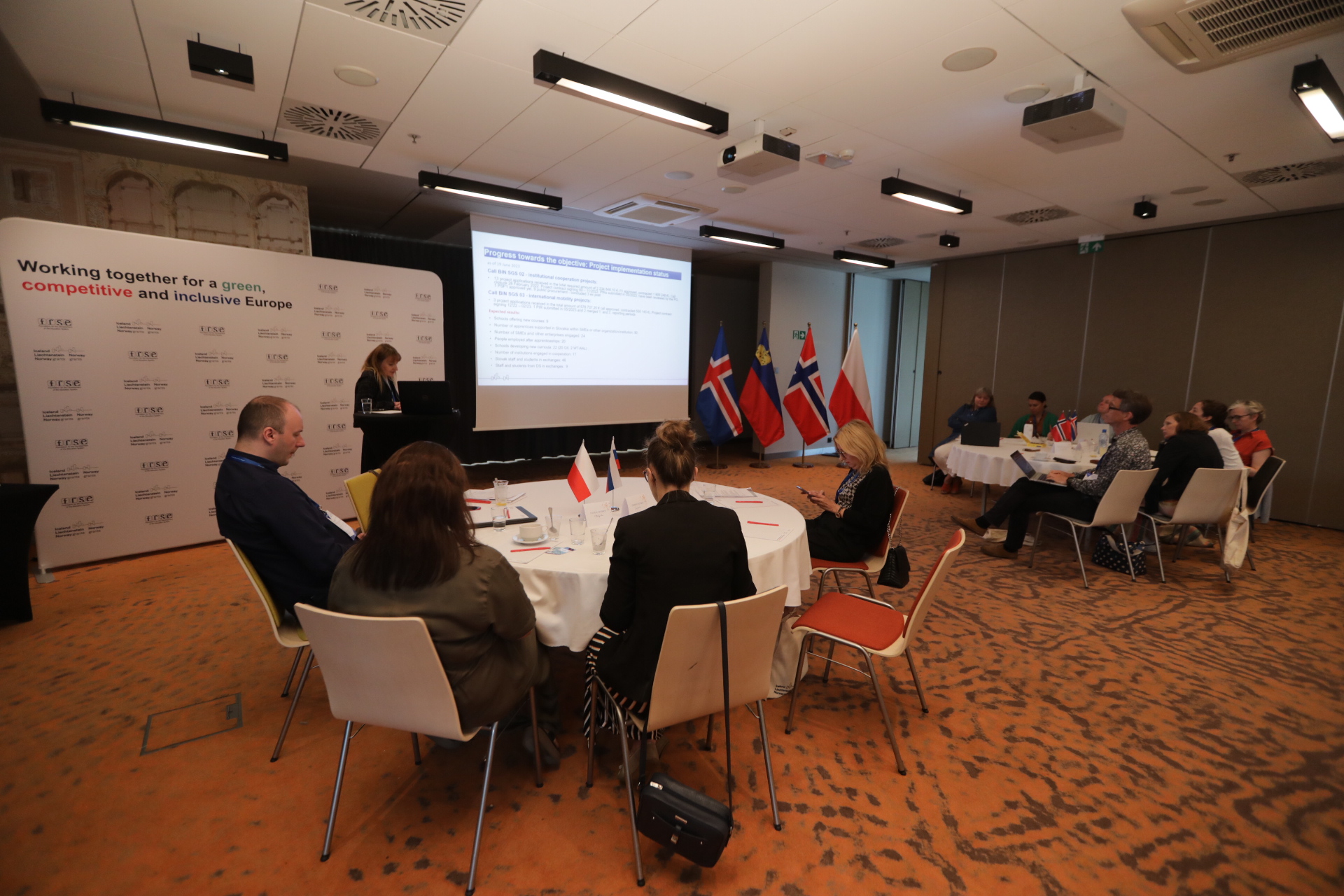 People at the tables, a person at the podium, a promotional stand, a projection of the presentation, the flags of the donor countries Iceland, Liechtenstein and Norway, and the flag of Poland.