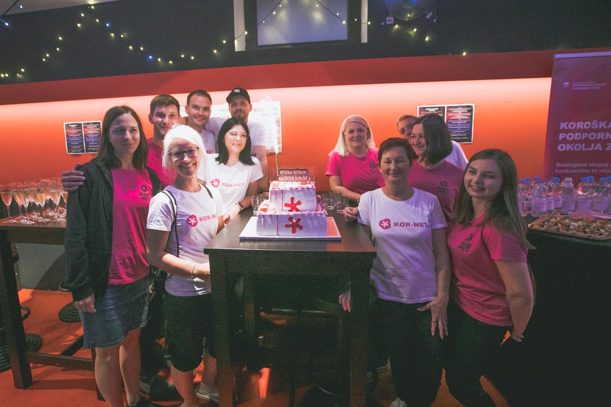 A group of people, one in white and one in pink T-shirts, gathered in a bar around a table with a two-storey square cake on it.