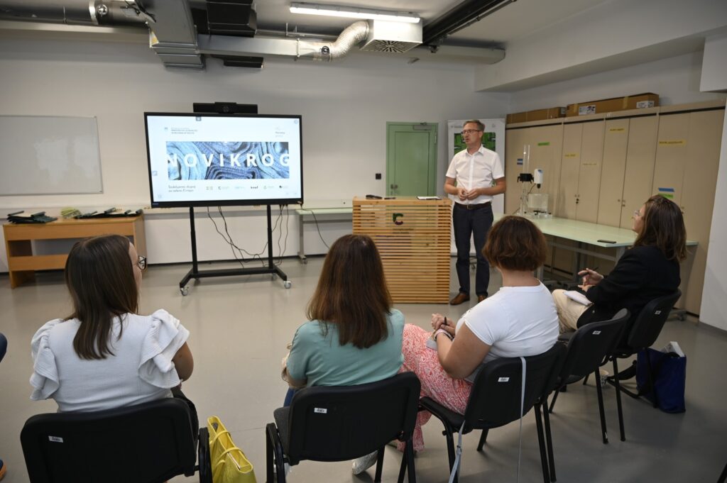 Screen with the acronym of the project NovIKroG, with a gentleman standing next to the speaker and the audience seated on chairs in front of him.