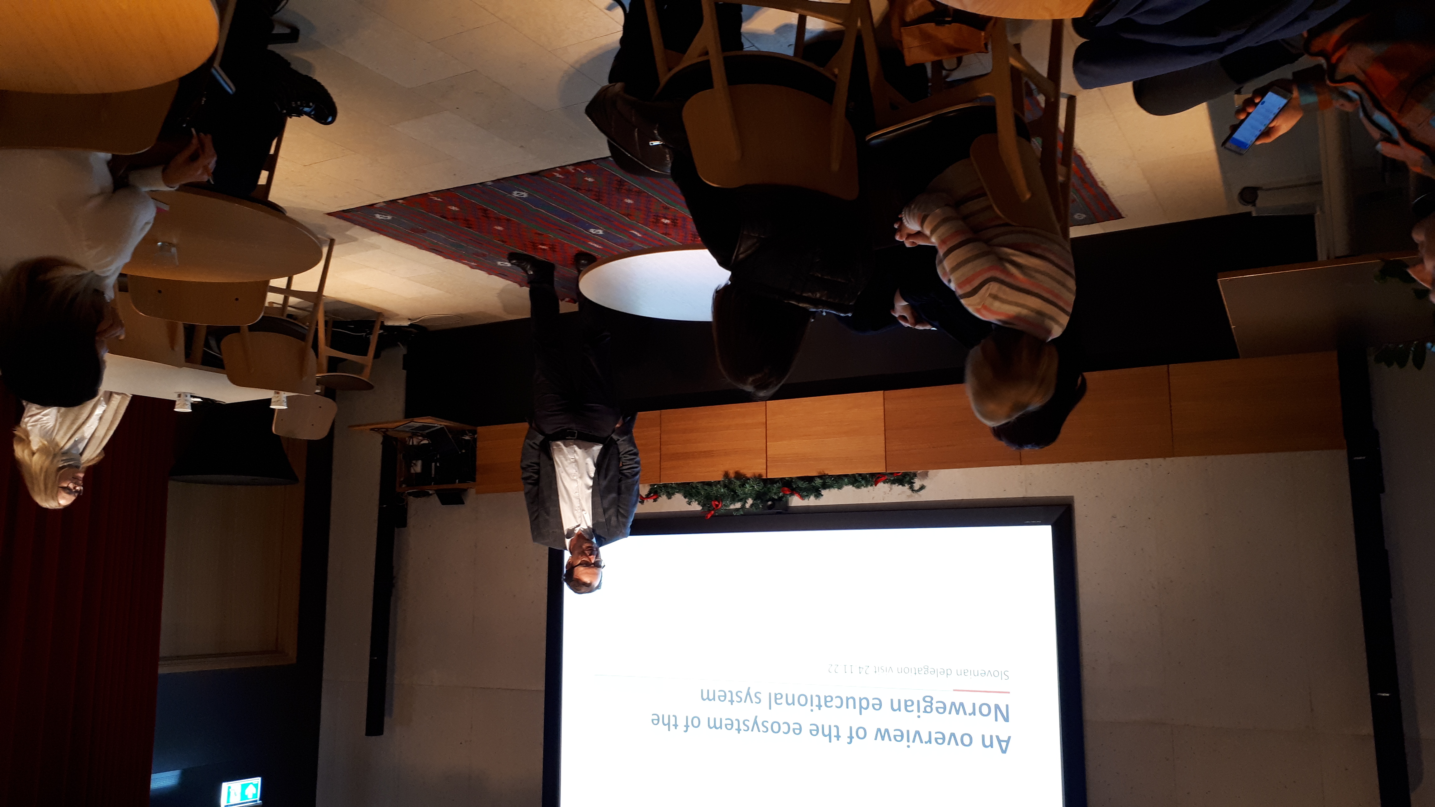 People gathered around a table of tables, the lecturer standing in front of a screen.