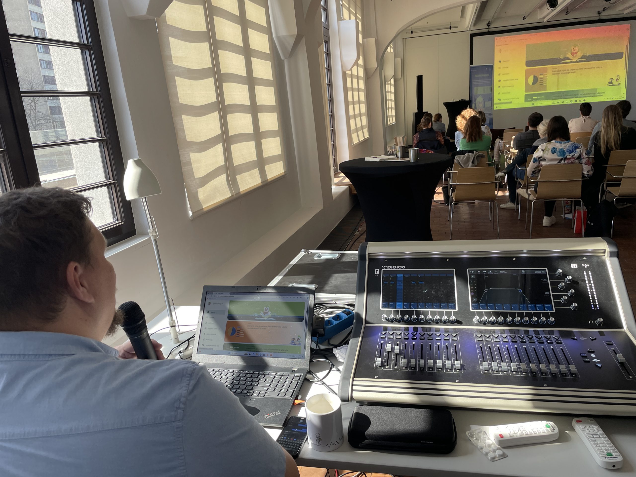 The technical equipment for the conference is set up in the back of the room, with the speaker sitting in a chair next to it, speaking into a microphone. In the foreground, participants are watching the screen.