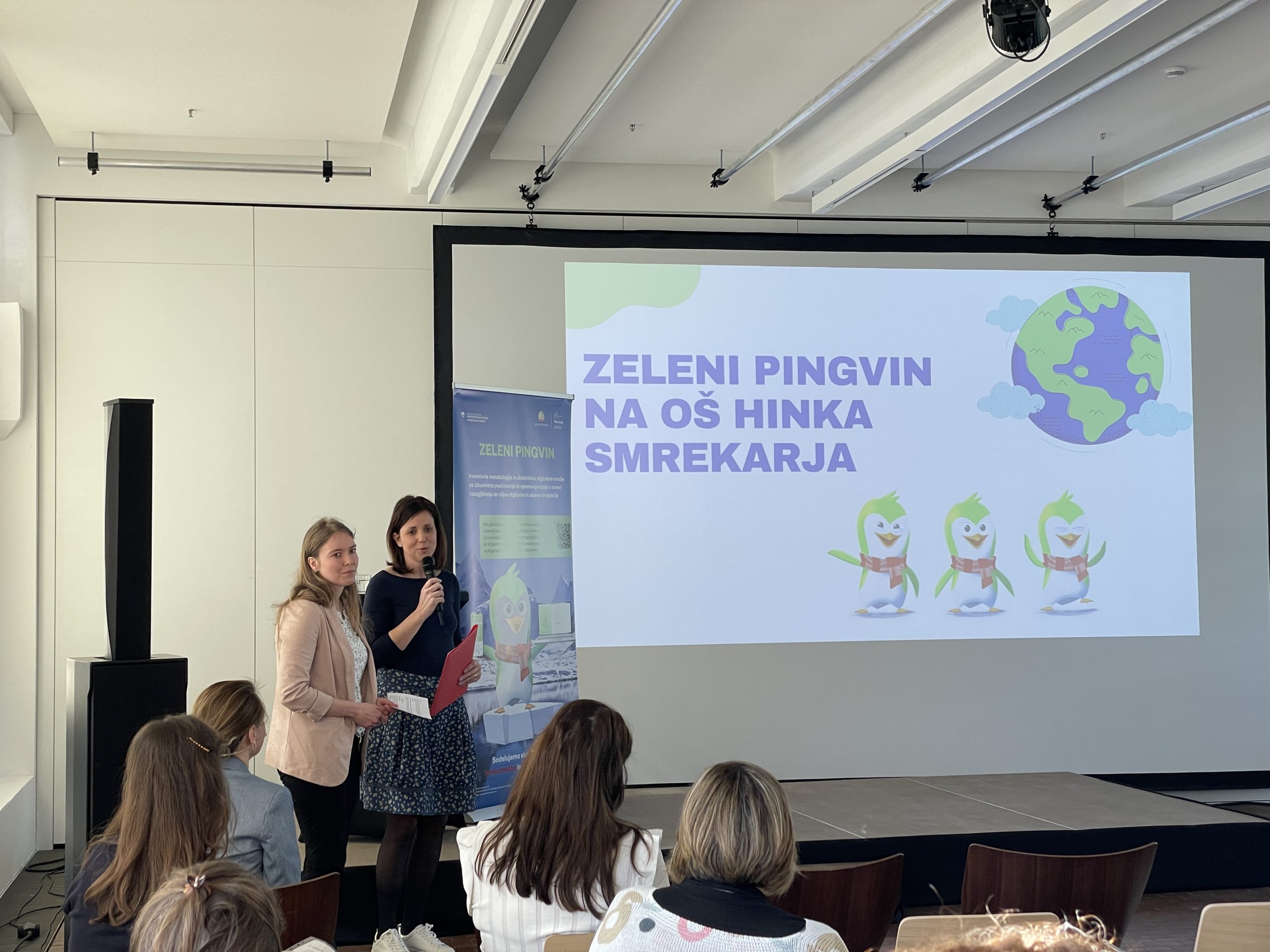 The speaker holds the microphone and speaks to the crowd. In the background, the screen displays a sign that reads Green Penguin at Hinko Smrekar Elementary School.