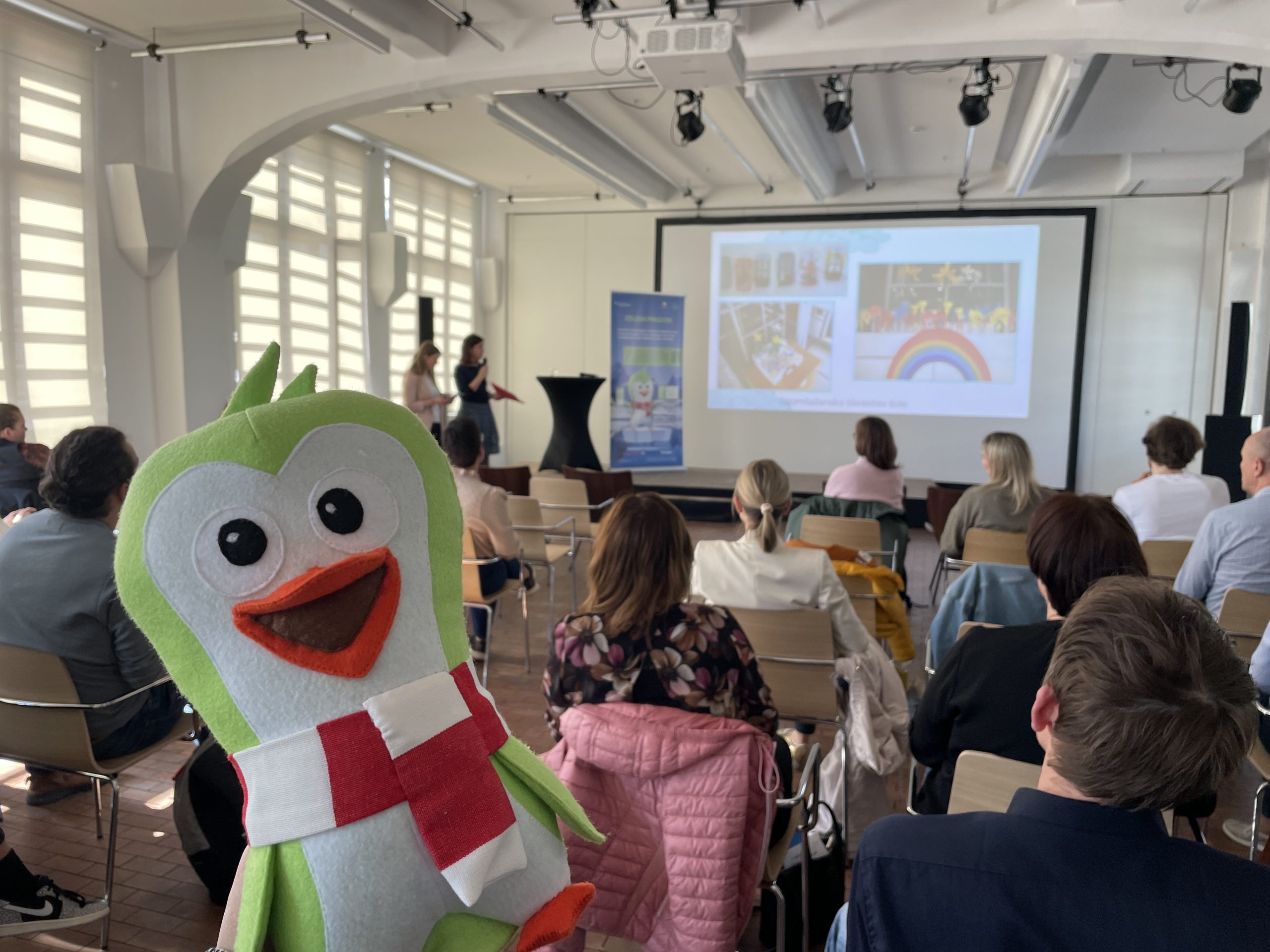 A plush Green Penguin mascot is placed in the centre, while participants watch the presentation on a screen in the background.