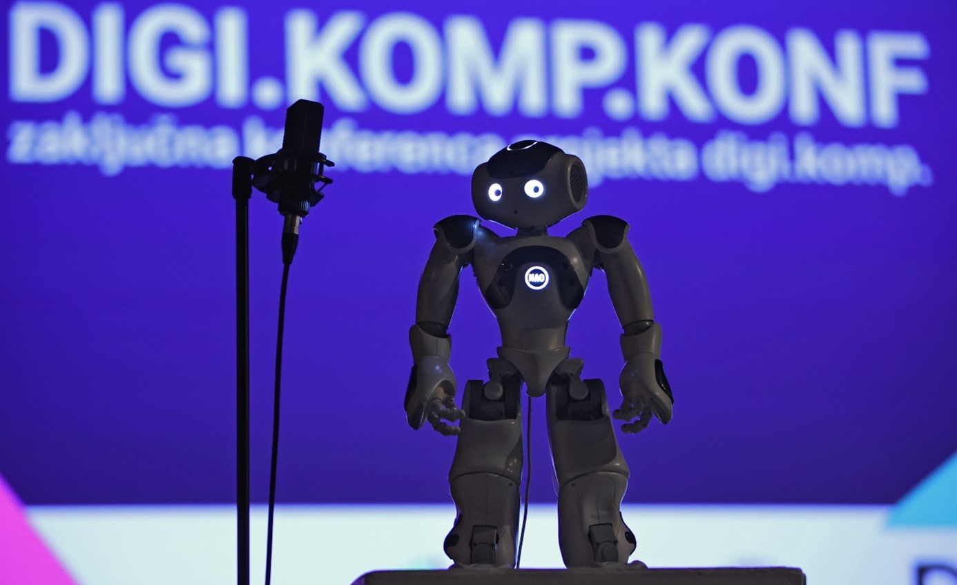 A robot stands on a stage with a microphone next to it. Behind the robot is a large screen with a presentation.