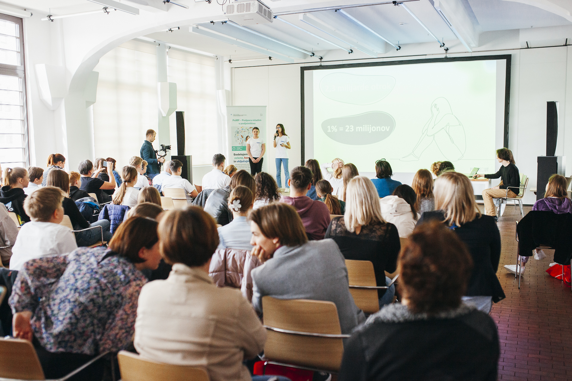 The audience sits and listens to a presentation by two young girls.